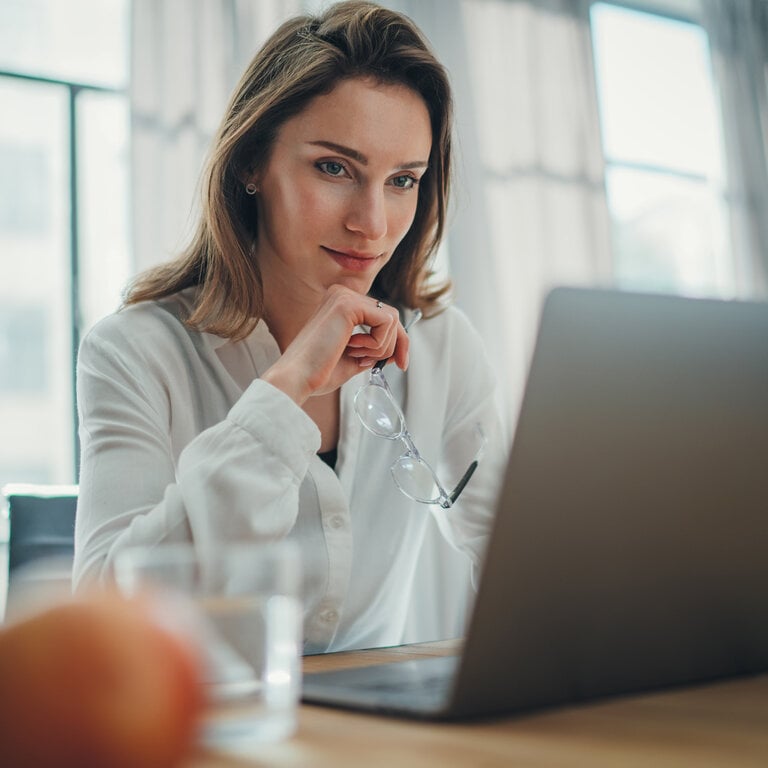 Eine Frau sitzt an einem Tisch vor einem Laptop, hält ihre Brille in der Hand und blickt konzentriert auf den Bildschirm.