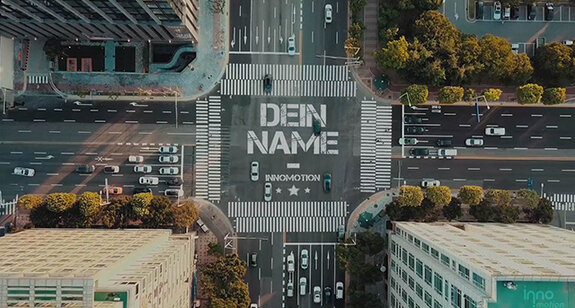 Luftaufnahme einer städtischen Kreuzung mit Schriftzug "Den Namen" auf der Straße, umgeben von modernen Gebäuden, Grünflächen und Fahrzeugen.