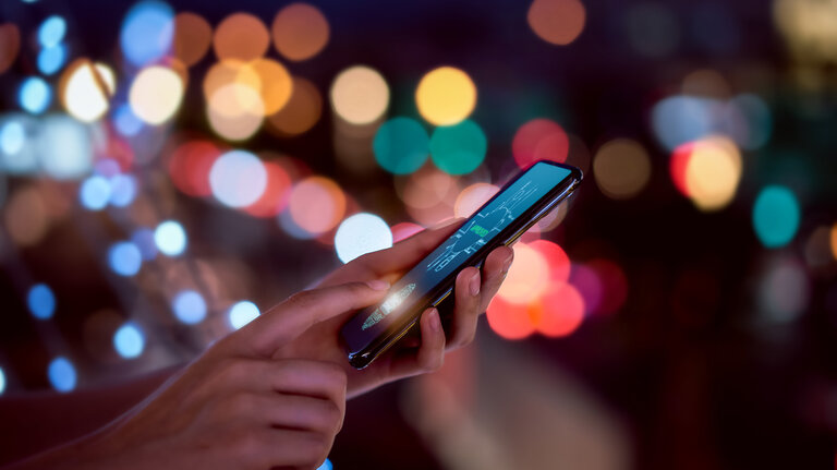 Nahaufnahme von Händen, die einen Fingerabdruck auf einem Smartphone scannen, um das Mobiltelefon zu entsperren – im Hintergrund verschwommene, farbige Lichter einer nächtlichen Stadt. Symbolbild für Sicherheit durch Identitäts-Technologie.