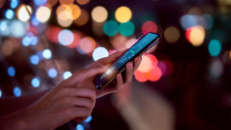 Nahaufnahme von Händen, die einen Fingerabdruck auf einem Smartphone scannen, um das Mobiltelefon zu entsperren – im Hintergrund verschwommene, farbige Lichter einer nächtlichen Stadt. Symbolbild für Sicherheit durch Identitäts-Technologie.