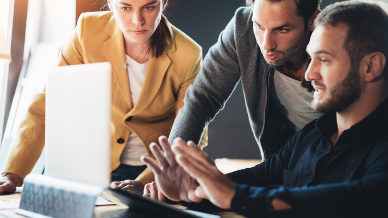 Das Bild zeigt drei Personen in einer professionellen Umgebung, die konzentriert an einem Laptop arbeiten. Eine Frau in einem gelben Blazer steht links und schaut aufmerksam auf den Bildschirm. Zwei Männer, einer sitzend und der andere stehend, scheinen in eine Diskussion vertieft zu sein, wobei der Sitzende seine Hände gestisch einsetzt, um etwas zu erklären. Die Szene vermittelt Teamarbeit und fokussiertes gemeinsames Arbeiten an einer Aufgabe oder einem Projekt.