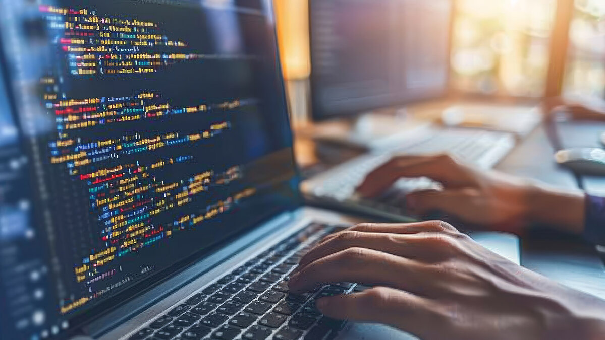 Person tippt Code auf einem Laptop, im Hintergrund weiterer Bildschirm mit Programmieroberfläche