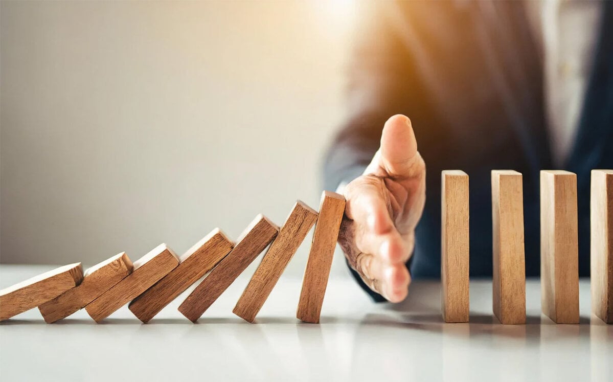 Eine Geschäftshand stoppt fallende Dominosteine – ein Symbol für die Vermeidung einer Unternehmenskrise und die Kontrolle potenzieller Störungen.