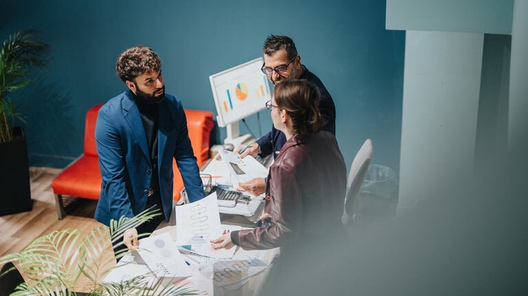 Drei Personen arbeiten gemeinsam in einem modernen Büro, analysieren Berichte und besprechen Erfolgsstrategien. Die Umgebung strahlt Professionalität, Zusammenarbeit und Konzentration aus – ideal für geschäftliche Zwecke.