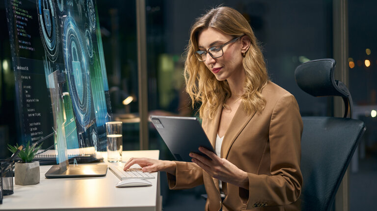 Fachfrau analysiert Daten auf einem Bildschirm und einem Tablet in einem modernen Arbeitsumfeld.