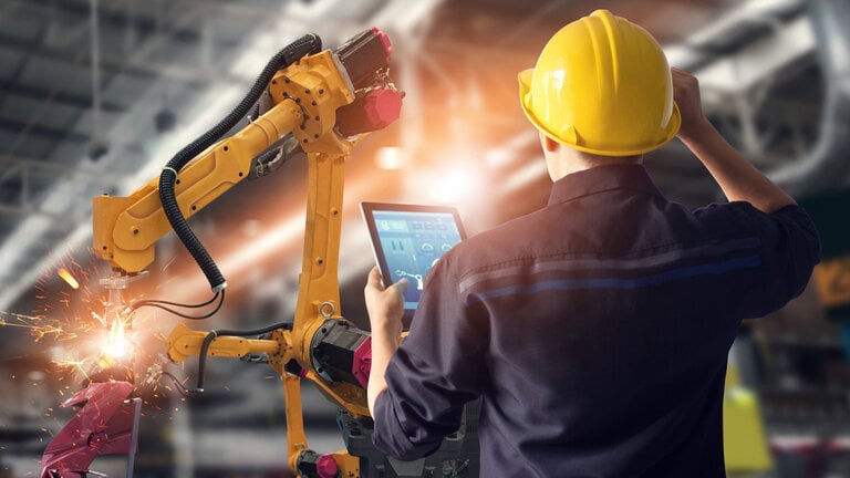 Ingenieur mit gelbem Schutzhelm bedient einen Tablet-Computer zur Steuerung eines industriellen Roboters, der in einer Fabrikhalle Funken sprüht.