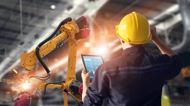 Ingenieur mit gelbem Schutzhelm bedient einen Tablet-Computer zur Steuerung eines industriellen Roboters, der in einer Fabrikhalle Funken sprüht.