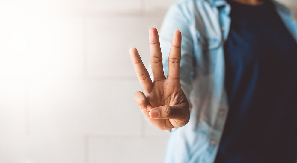 Hand, die drei Finger hebt, symbolisch für eine Zahl oder ein Konzept, vor einem hellen und neutralen Hintergrund.