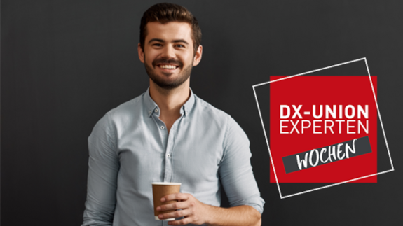 Ein Mann mit Bart hält eine Kaffeetasse und lächelt, während im Hintergrund ein schwarzer Hintergrund mit dem Text "DX-Union Experten Wochen" zu sehen ist.
