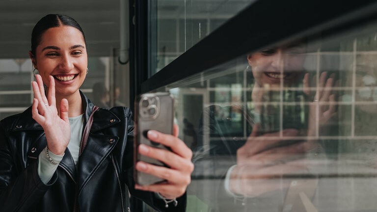 Zwei junge Frauen stehen dicht beieinander, machen ein Selfie und winken in die Kamera ihres Smartphones. Ihre Spiegelbilder sind im Glas zu sehen, während sie lächeln und in die Kamera winken.