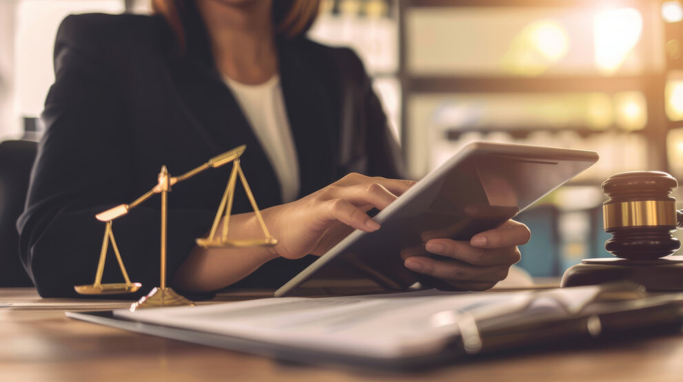 Eine Person in einem formellen Anzug benutzt ein Tablet an einem Schreibtisch, der mit einer Waage, einem Hammer und Dokumenten geschmückt ist, was auf ein juristisches oder gerichtliches Umfeld hinweist.
