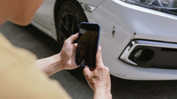 Ein junger Mann hält ein Smartphone in der Hand und fotografiert sein nach einem Unfall verbeultes Auto. Ein Versicherungsagent mit einem Klemmbrett begutachtet den Schaden und erstellt einen Schadensbericht.
