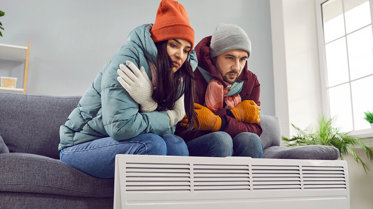 Menschen frieren und wärmen sich zu Hause neben einem Elektroheizgerät. Verärgerte, warm angezogene Ehepartner sitzen auf dem Sofa, frieren und versuchen sich aufzuwärmen. Heizungsprobleme, Konzept der Energiekrise