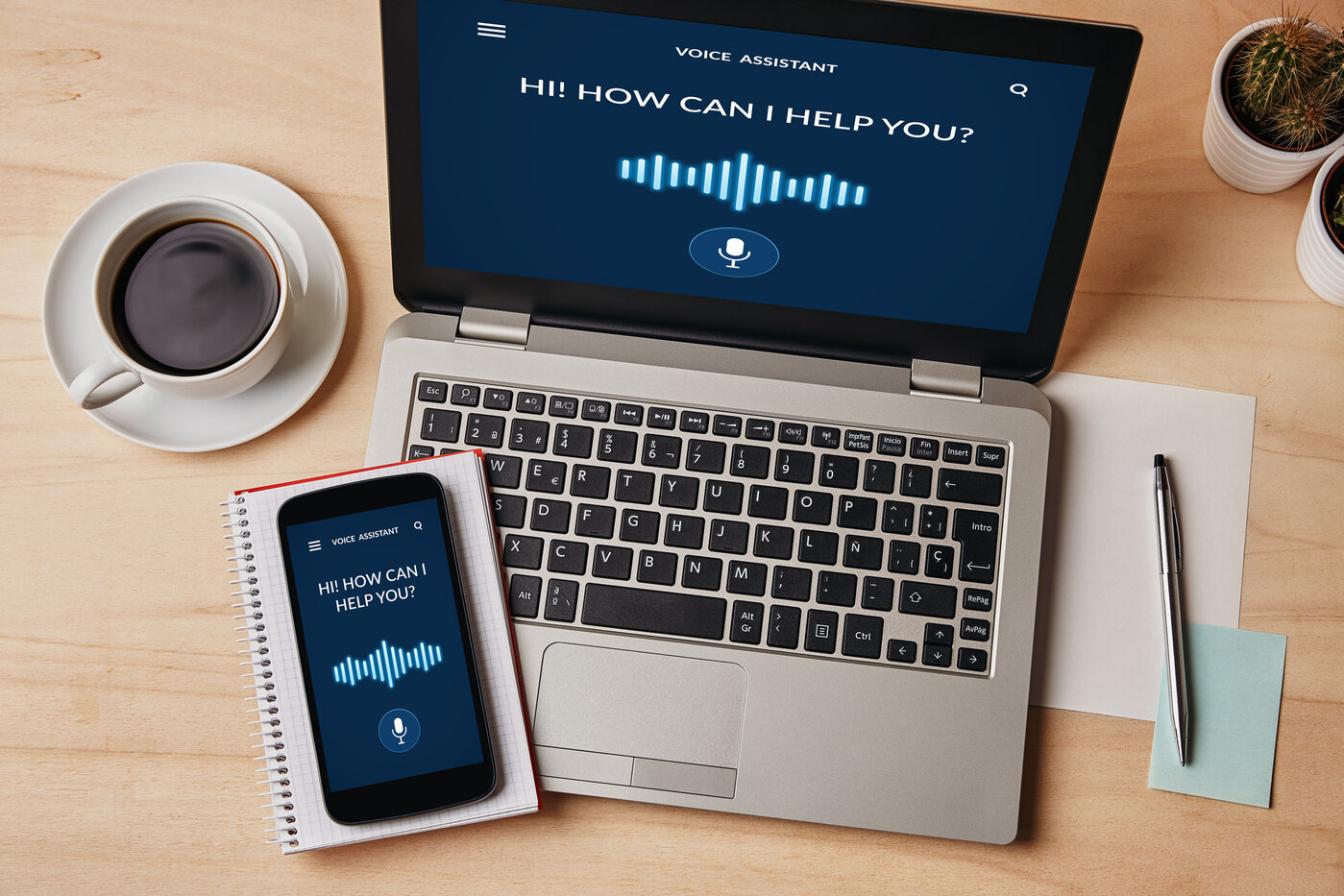 Ein Laptop und ein Smartphone mit einer aktiven Sprachassistenten-App, die die Nachricht "Hi! How can I help you?" anzeigt. Die Geräte liegen auf einem Schreibtisch, begleitet von einer Tasse Kaffee, einem Notizblock und einigen Büroartikeln.