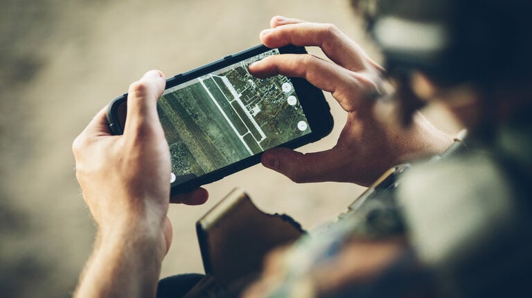 Eine Person betrachtet eine Satellitenkarte auf einem Smartphone, möglicherweise in einer taktischen oder militärischen Umgebung, basierend auf der Kleidung und dem Hintergrund.