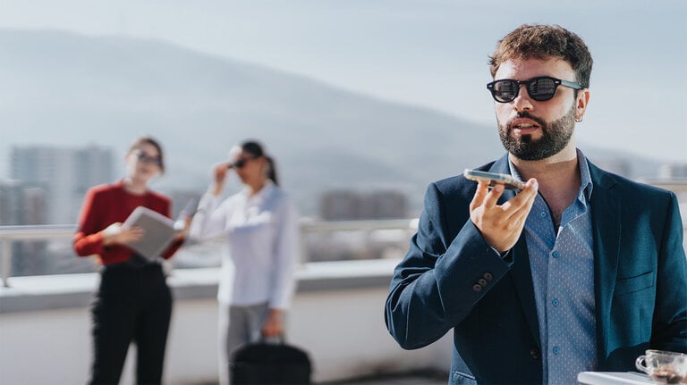 Ein selbstbewusster Geschäftsmann mit Sonnenbrille nutzt auf einem Dach vor einer städtischen Kulisse den Sprachassistenten seines Smartphones. Im Hintergrund unterhalten sich zwei verschwommene Personen, was eine lebhafte Geschäftsszene schafft.