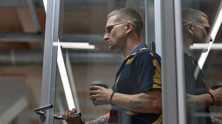 Seitenansicht eines modernen Geschäftsmannes in einer Türöffnung, der mit einem wiederverwendbaren Kaffeebecher die Tür öffnet, während er das Büro in einem Coworking Space verlässt, Bildausschnitt