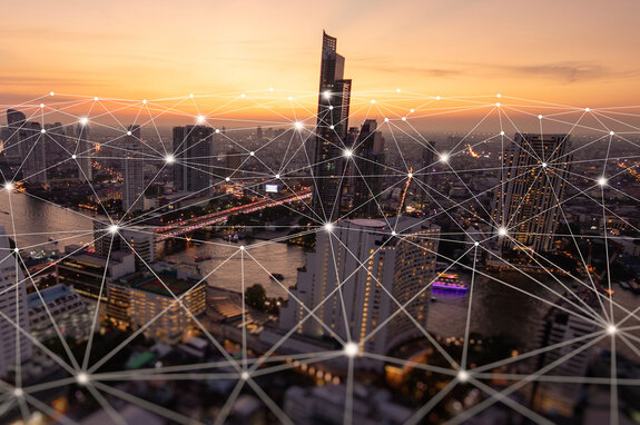 Eine Stadtansicht bei Sonnenuntergang, verbunden durch ein digitales Netzwerk mit leuchtenden Knotenpunkten, das die digitale Vernetzung und Smart-City-Konzepte symbolisiert.