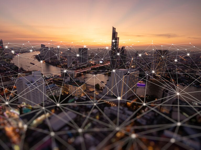 Eine Stadtansicht bei Sonnenuntergang, verbunden durch ein digitales Netzwerk mit leuchtenden Knotenpunkten, das die digitale Vernetzung und Smart-City-Konzepte symbolisiert.