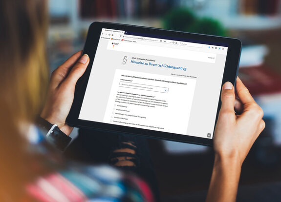 Eine Person hält ein Tablet, auf dem eine Website mit dem Titel 'Hinweise zu Ihrem Schlichtungsantrag' angezeigt wird.