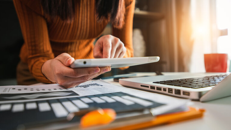 Eine Person arbeitet mit einem Tablet und Laptop auf einem Schreibtisch, umgeben von Dokumenten und Diagrammen, während Sonnenlicht durch das Fenster fällt.