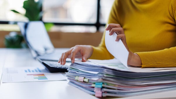 Eine Geschäftsfrau arbeitet an einem überfüllten Schreibtisch im Büro und durchsucht Stapel von Papierakten, um unvollständige Dokumente in Ordnern zu finden