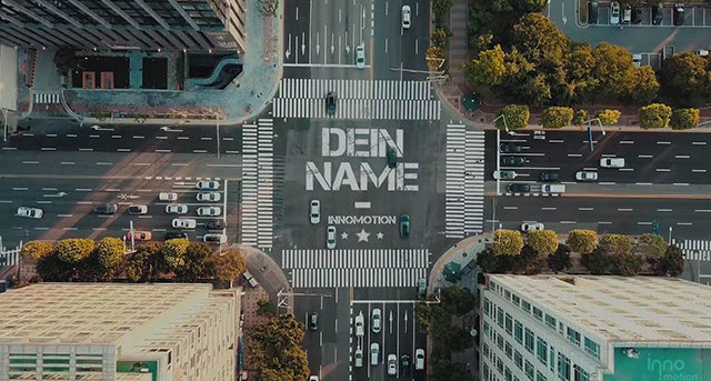 Luftaufnahme einer städtischen Kreuzung mit Schriftzug "Den Namen" auf der Straße, umgeben von modernen Gebäuden, Grünflächen und Fahrzeugen.
