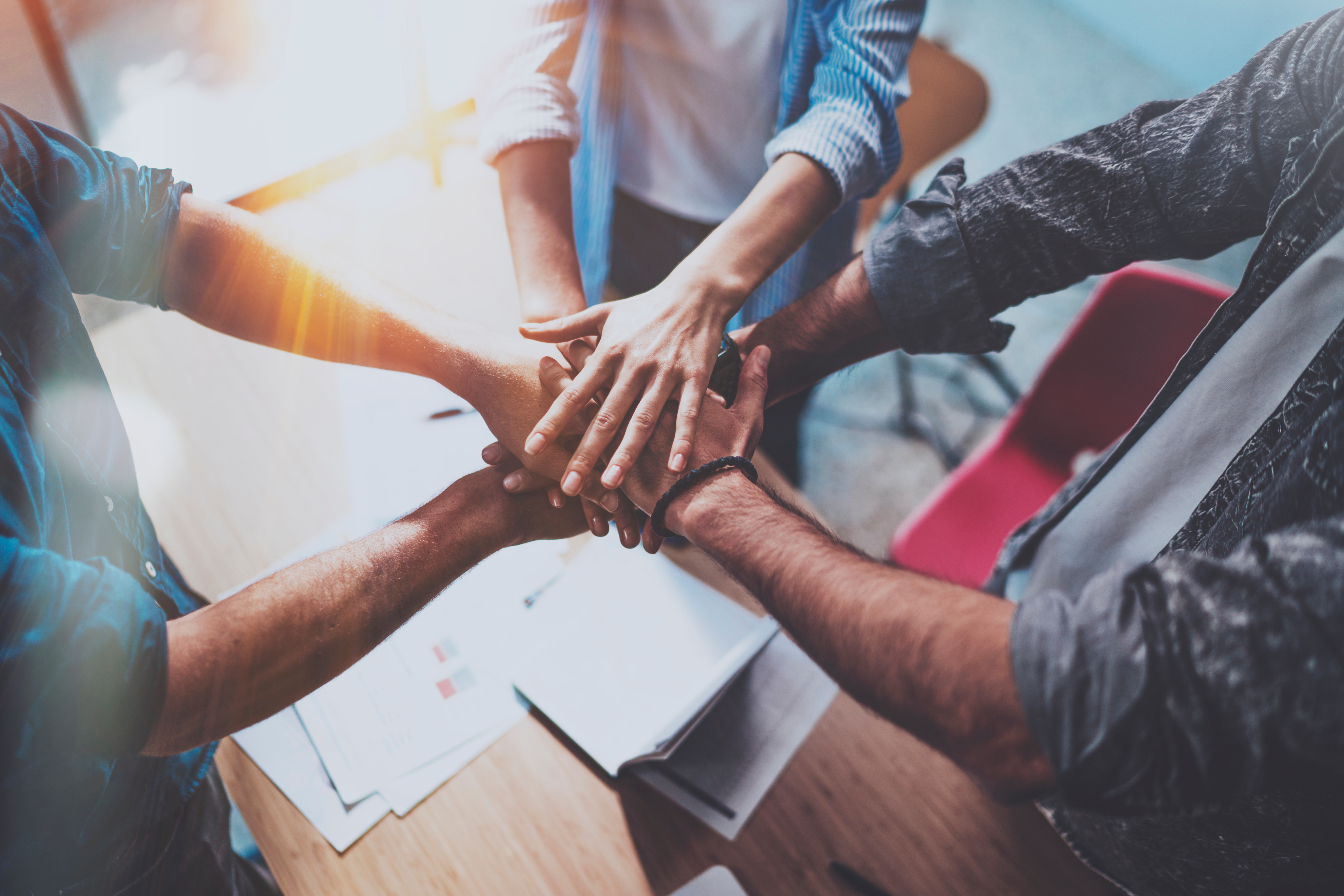 Teammitglieder legen ihre Hände übereinander, um Zusammenarbeit und Gemeinschaft zu symbolisieren, mit Dokumenten und Sonnenlicht im Hintergrund.