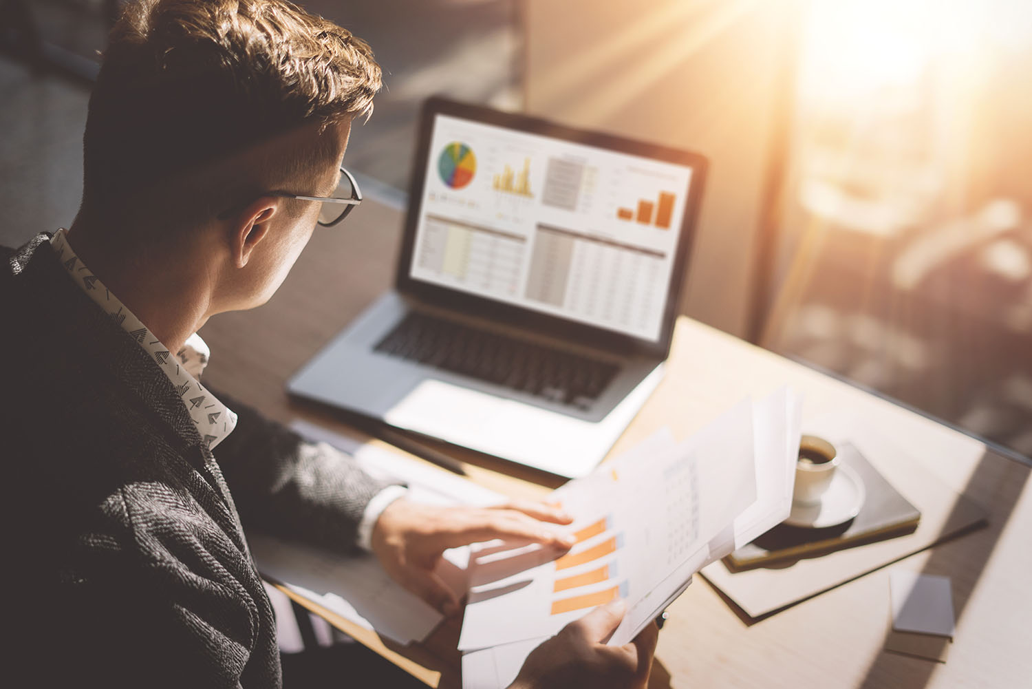 Junger Finanzmarktanalyst mit Brille, der in einem sonnigen Büro an einem Laptop arbeitet, während er an einem Holztisch sitzt. Geschäftsmann analysiert Dokument.