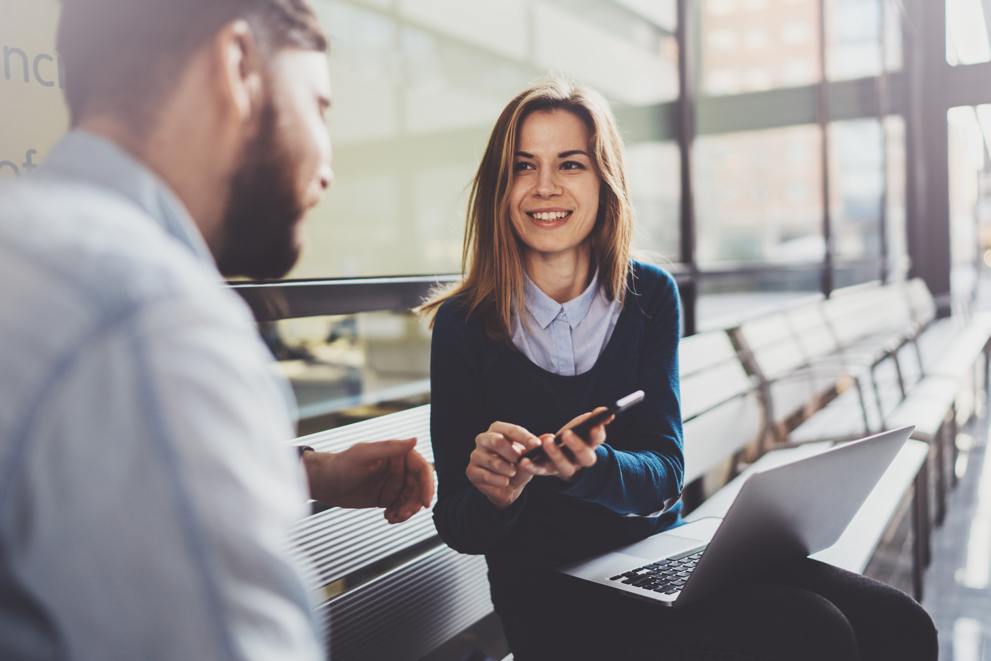 Zwei Personen sitzen auf einer Bank in einem modernen Gebäude, eine Person hält ein Smartphone und lächelt, während ein Laptop neben ihnen liegt.