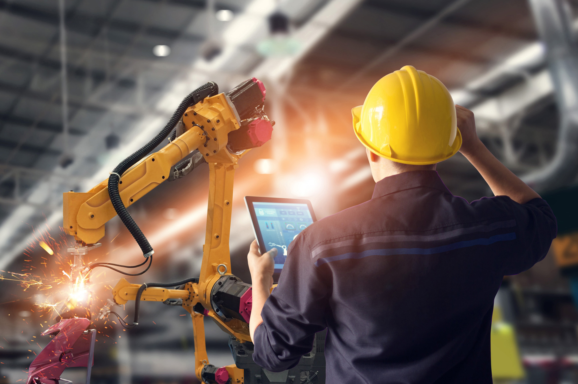 Ingenieur mit gelbem Schutzhelm bedient einen Tablet-Computer zur Steuerung eines industriellen Roboters, der in einer Fabrikhalle Funken sprüht.