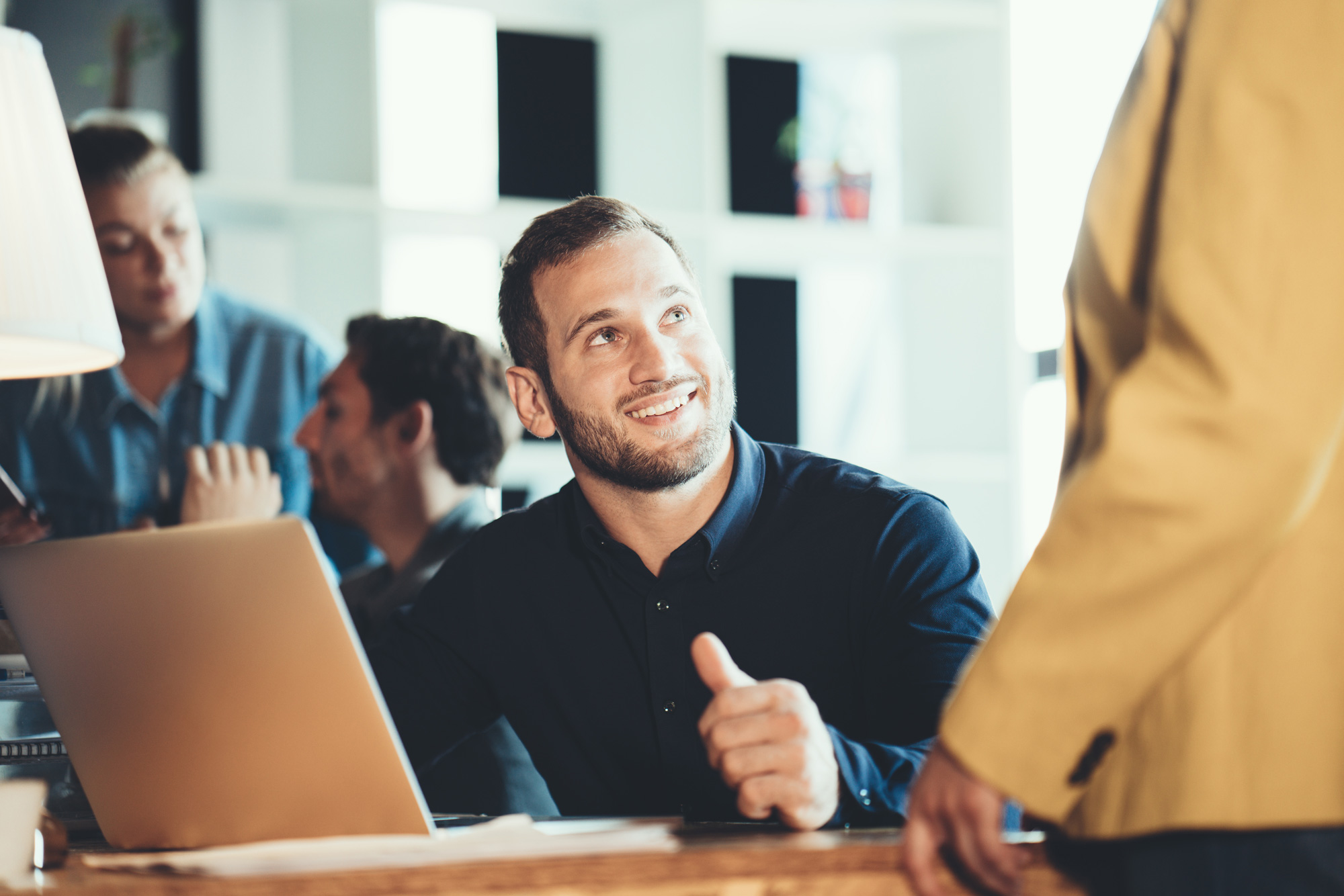 Ein lächelnder Mitarbeiter in einem modernen Büro interagiert mit einer Kolleg:in, umgeben von weiteren Kolleg:innen im Hintergrund und einem Laptop auf dem Schreibtisch.