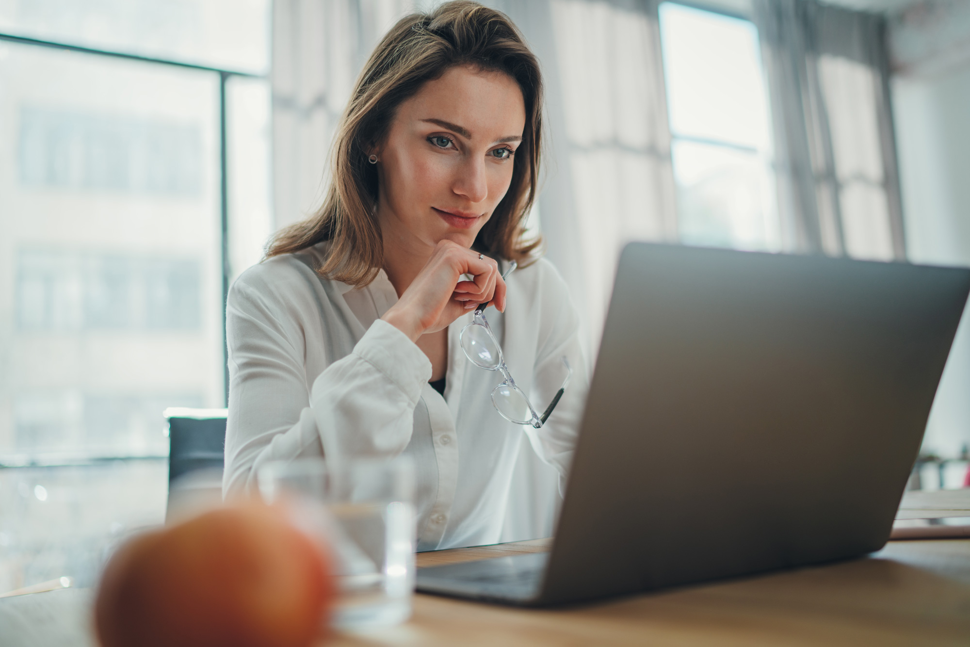 Eine Frau sitzt an einem Tisch vor einem Laptop, hält ihre Brille in der Hand und blickt konzentriert auf den Bildschirm.