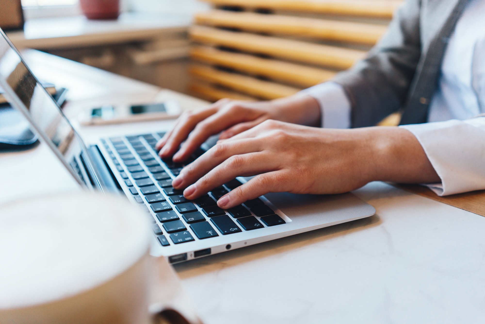 Hände einer Person, die auf der Tastatur eines Laptops tippt, mit verschwommenem Getränk im Vordergrund.
